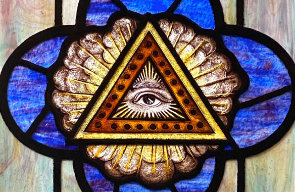 Close-up of Masonic eye in triangle surrounded by rays on blue 4-leafed shape, stained glass window.